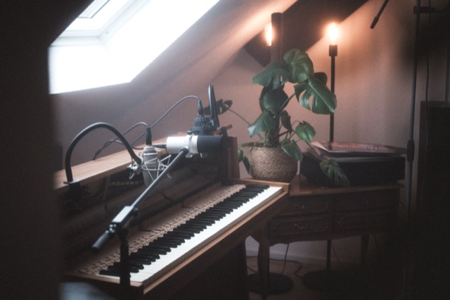 Großmembranmikrofon vor dem Upright Piano unter der Dachschräge, daneben eine Monstera.