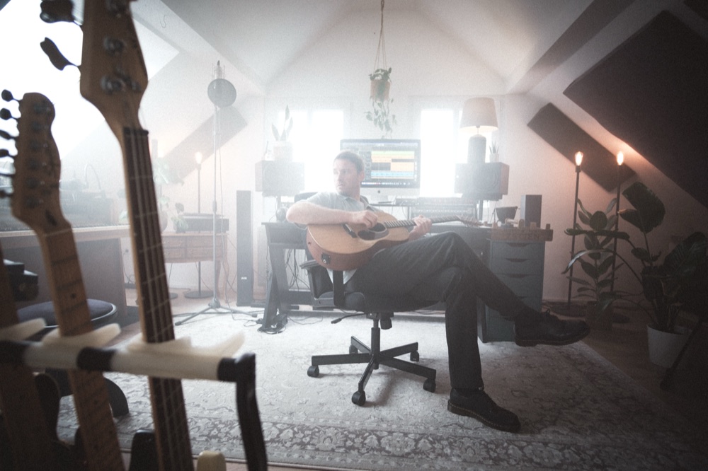 Phil Young sitzt mit Westerngitarre am Schreibtisch, im Vordergrund unscharf mehrere Gitarren im Ständer.